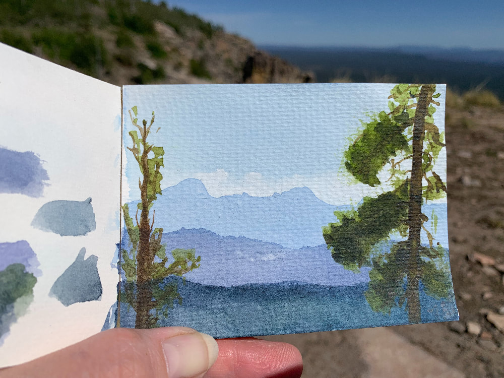 A photo of a hand holding open a small sketchbook. The right page of the sketchbook has a watercolor painting of a view looking out from the Mogollon Rim. The painting shows two trees on the edge of the rim and the mountains receding in shades of blue into the distance. There's a soft blue sky and white clouds. A portion of the left page of the sketchbook shows swatches of watercolor. Behind the sketchbook, the background is soft and there’s a rocky ground with greenery on the top left and blue sky on the top right. 